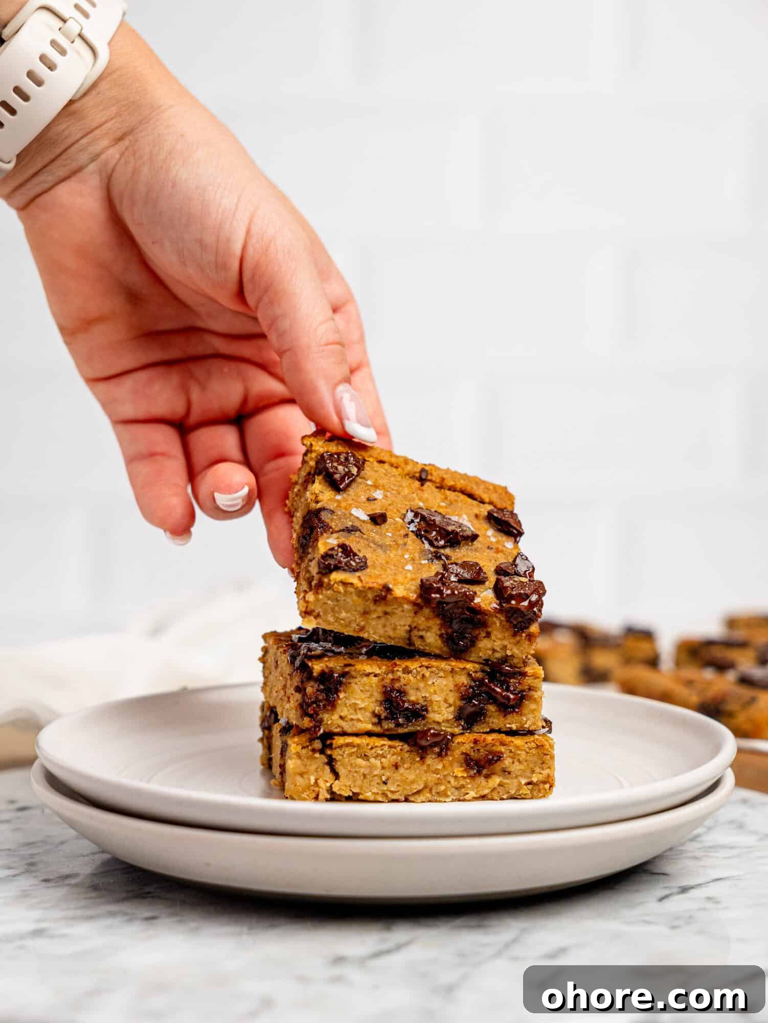 Gooey Chickpea Cookie Bars — Protein-Packed Dessert Bars 8 Three chickpea cookie bars stacked on top of each other on a white plate, a hand is picking up the one on top.