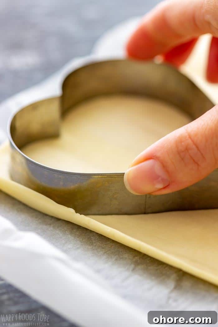 Flaky Puff Pastry Hearts for Valentine’s Day Gifts 3 A hand is pressing a heart-shaped cookie cutter into a sheet of puff pastry on a parchment-lined surface.