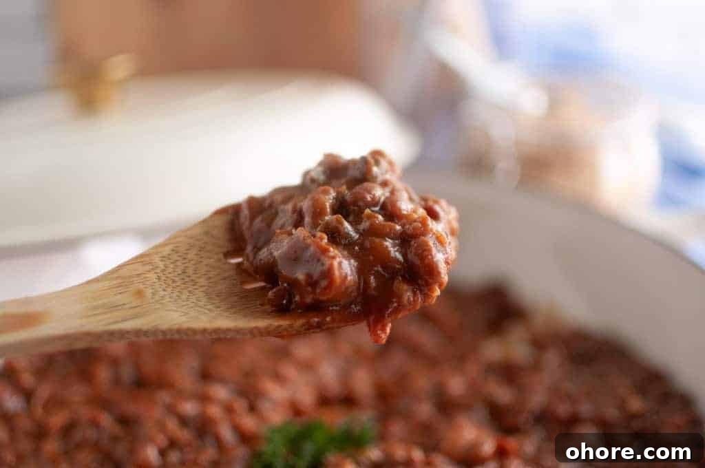 A wooden spoon holds a serving of Baked Bean Casserole above a pot filled with more bean casserole.
