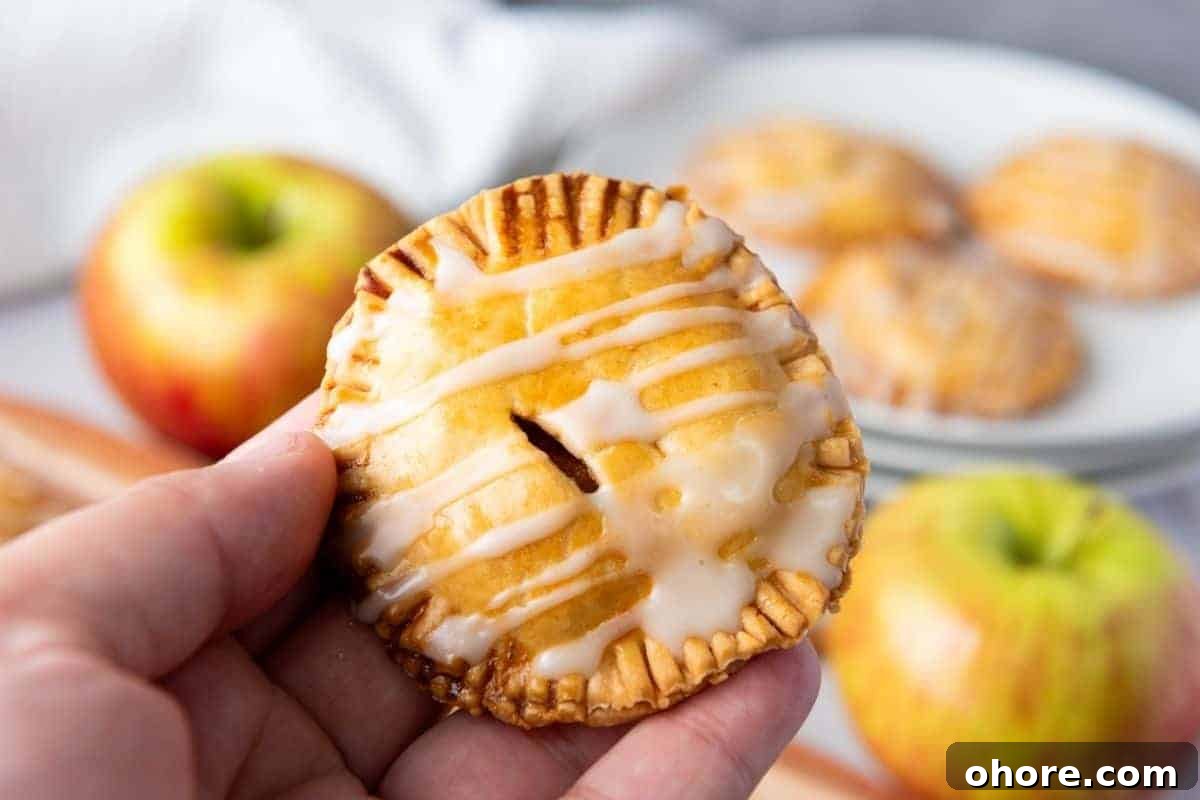A hand holds a small round hand pie with icing drizzle; apples and more hand pies are visible in the background.