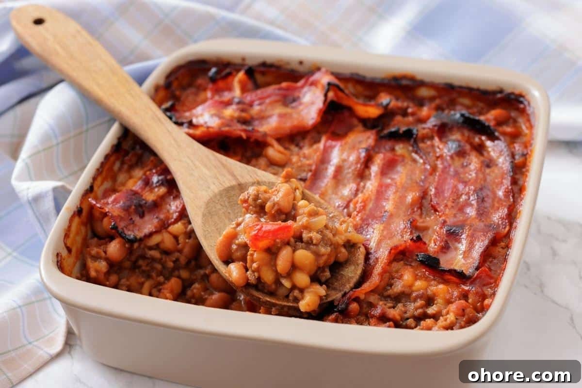 A square baking dish filled with Southern Ground Beef & Baked Bean Casserole and topped with strips of cooked bacon; a wooden spoon holds a serving.