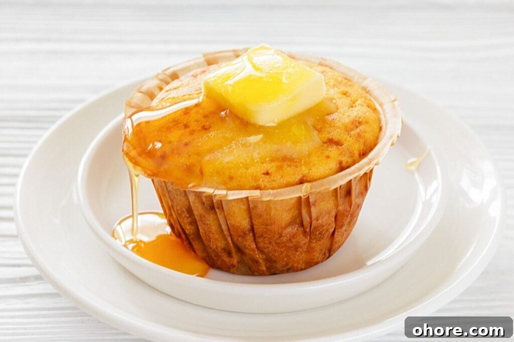 A Honey Cornbread muffin on a white plate, topped with a pat of butter and drizzled with syrup.