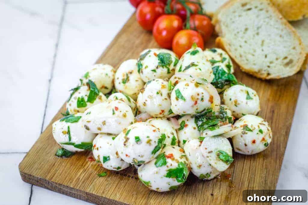 A pile of herb-marinated mozzarella balls with herbs on a wooden board, next to sliced bread and a bunch of cherry tomatoes.