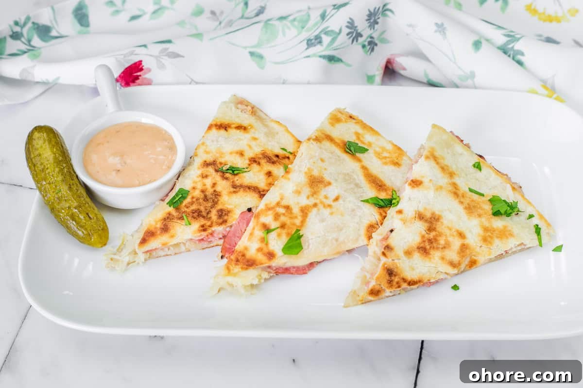 Three Reuben quesadilla slices with herbs on a white plate, accompanied by a pickle and a small dish of dipping sauce.