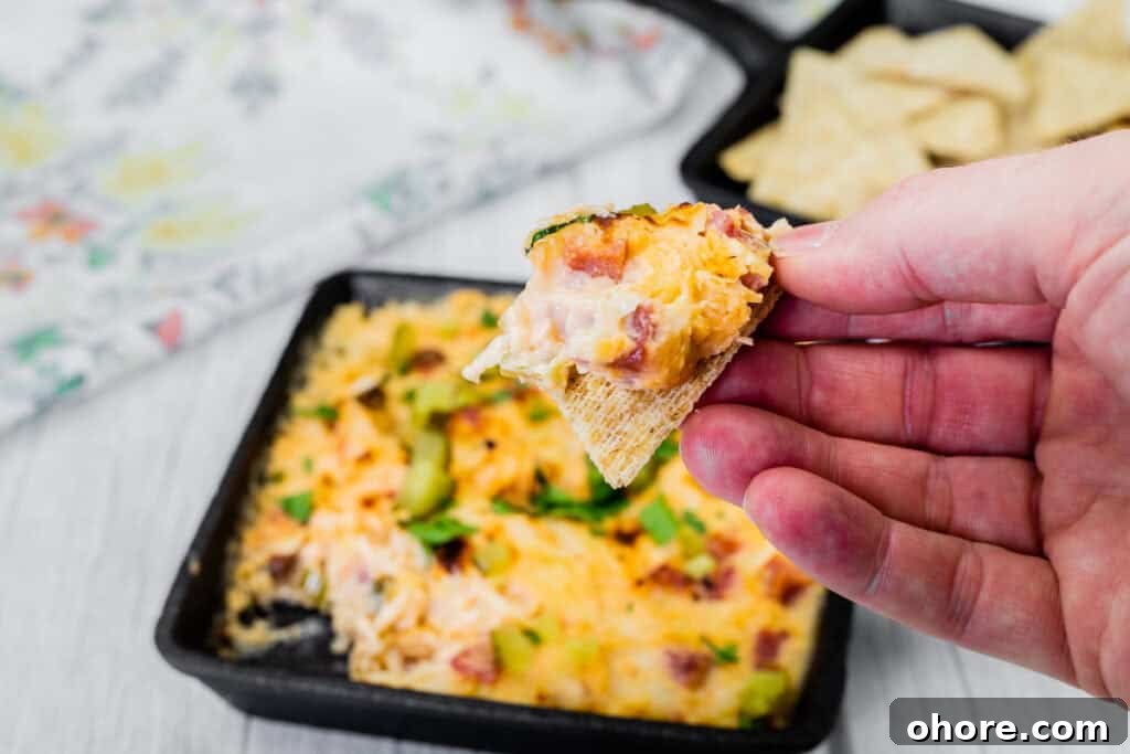 A hand holds a chip with Hot Reuben Dip from a skillet of the dip and cracker.