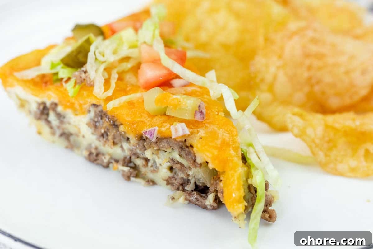 A slice of cheeseburger pie topped with shredded lettuce, diced tomato, onions, and pickles, served with potato chips on a white plate.