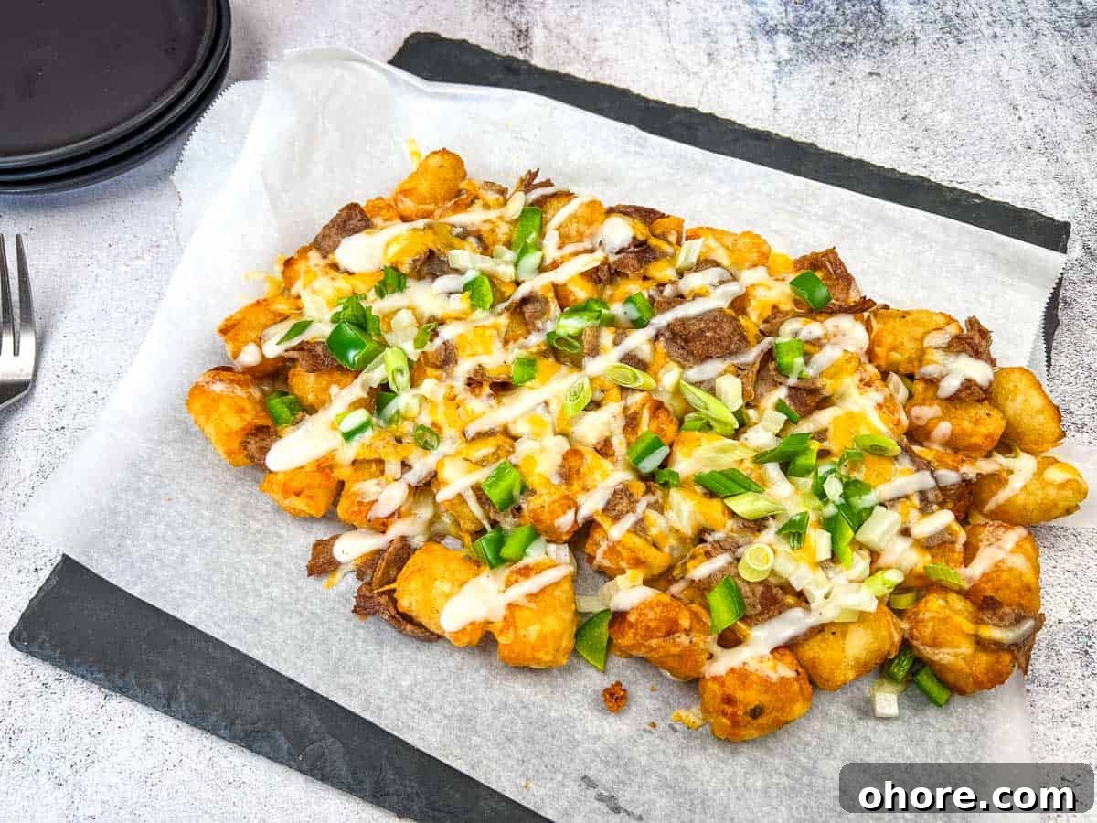 Philly Cheesesteak Loaded Tots on parchment paper, topped with Alfredo cheese, green onions, and a drizzle of white sauce, placed on a dark rectangular slate plate.