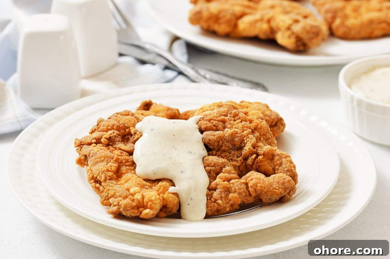 A plate of fried chicken steak topped with white gravy, with more fried pieces and a bowl of gravy in the background.
