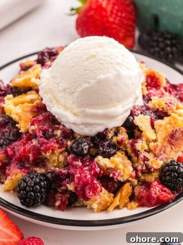 Berry cobbler with cake mix topped with vanilla ice cream on a plate with fresh berries nearby.