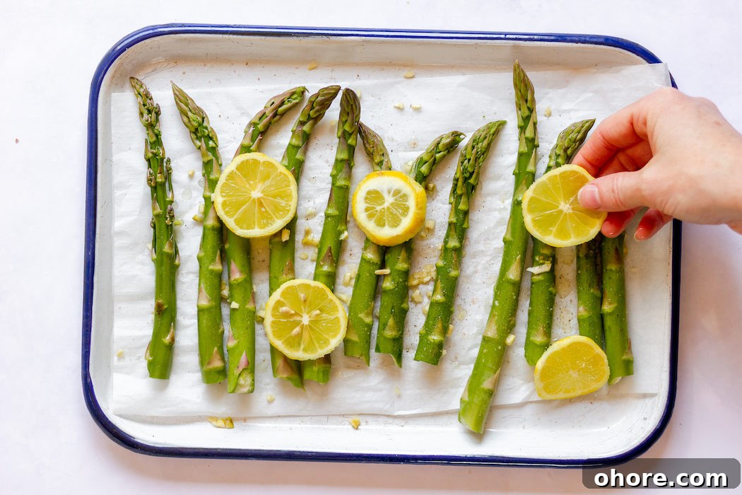 Image, taken from above, shows a hand placing lemon slices on top of asparagus stalks and garlic.