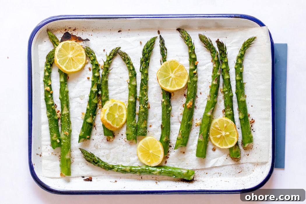 Image, taken from above shows the asparagus stalks on a tray with lemon and garlic, ready to be roasted. 