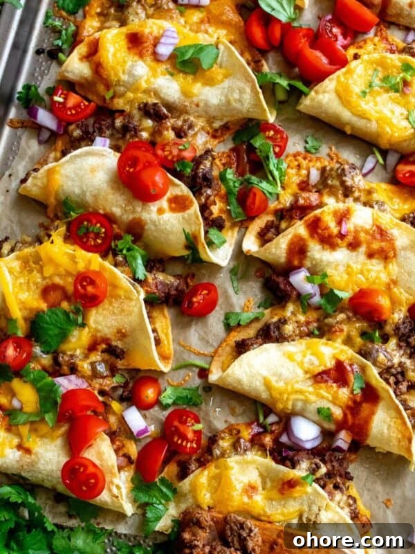 Crispy Beef Tacos on parchment liked baking sheet with hot sauce and tomatoes.