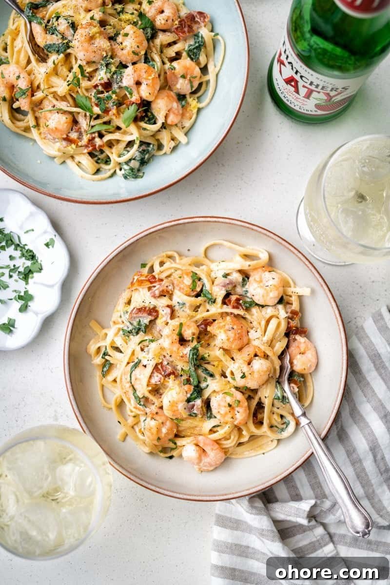 Two bowls of creamy Tuscan shrimp pasta with spinach and sun-dried tomatoes sit on a white table beside a glass of ice, chopped herbs on a small plate, a striped napkin, and a bottle of sparkling water.