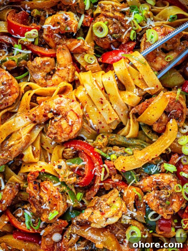 A close-up of rasta pasta with jerk shrimp with wide noodles, shrimp, sliced red peppers, green onions, and a sprinkle of red pepper flakes, garnished with grated cheese and herbs.