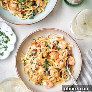 Two bowls of creamy Tuscan shrimp pasta with spinach and sun-dried tomatoes sit on a white table beside a glass of ice, chopped herbs on a small plate, a striped napkin, and a bottle of sparkling water.