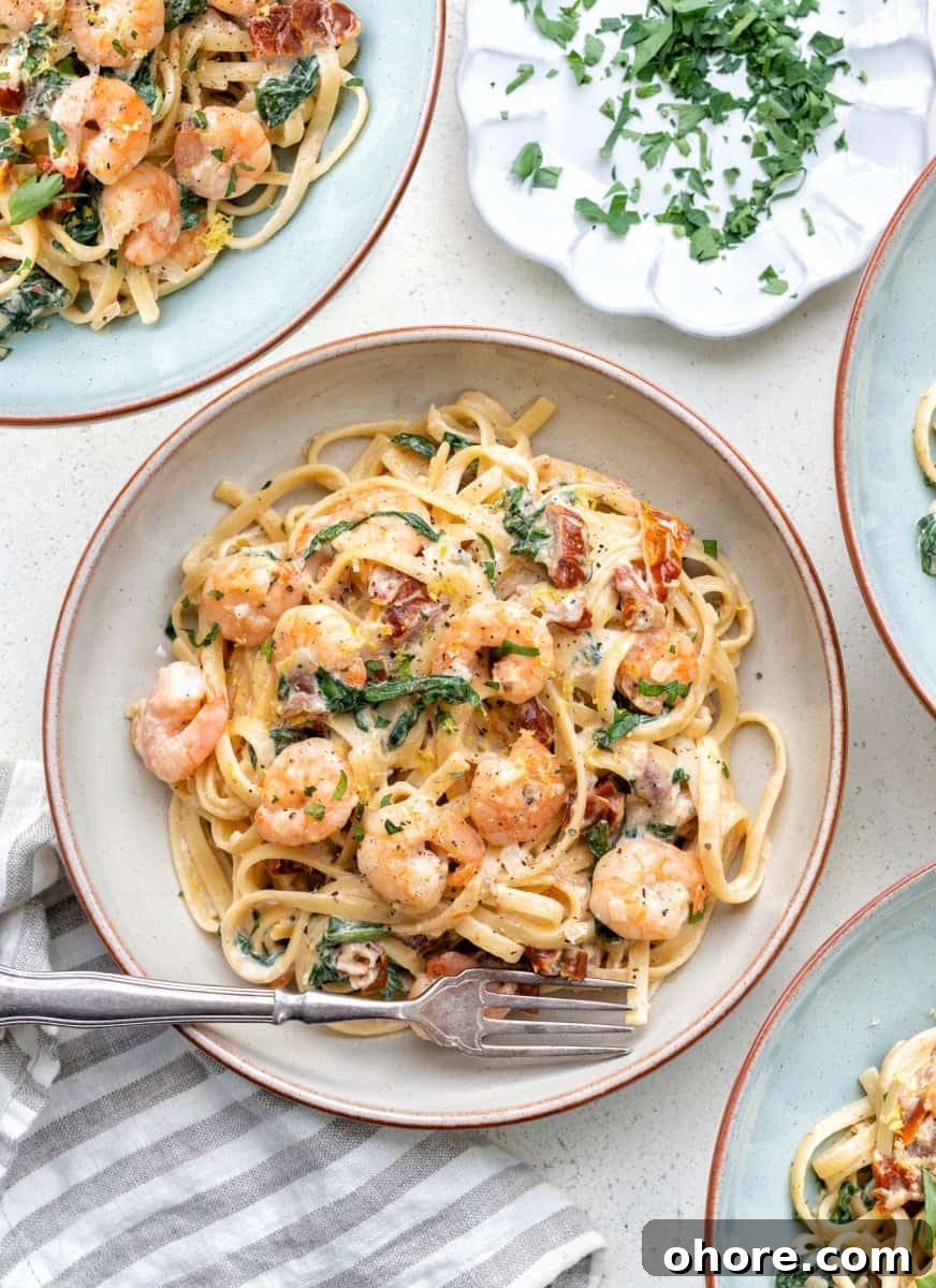 A bowl of creamy Tuscan shrimp pasta with sun-dried tomatoes, spinach, and herbs, garnished with grated cheese, sits on a plate with a fork. Other similar plates and a dish of chopped parsley are nearby.