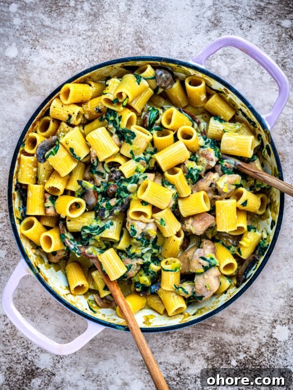 A large pot filled with rigatoni pasta, creamy sauce, mushrooms, diced chicken, fresh spinach, and parmesan. Two wooden spoons rest inside the pot on a textured surface.