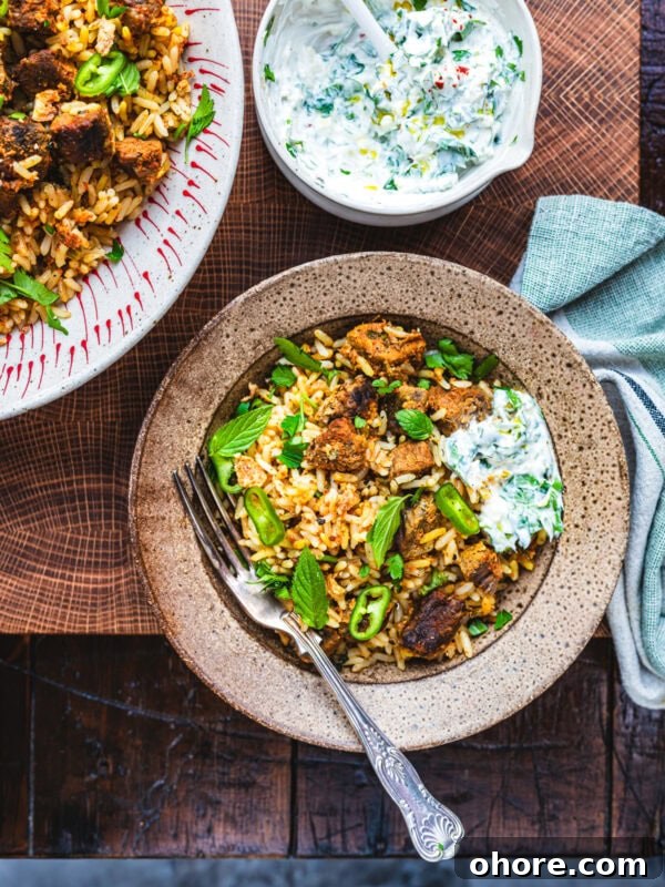 A bowl of spiced beef biryani with chunks of meat, garnished with fresh mint and sliced green chili, served with a portion of raita. A fork rests in the bowl, and a bowl of raita is nearby on a wooden surface.