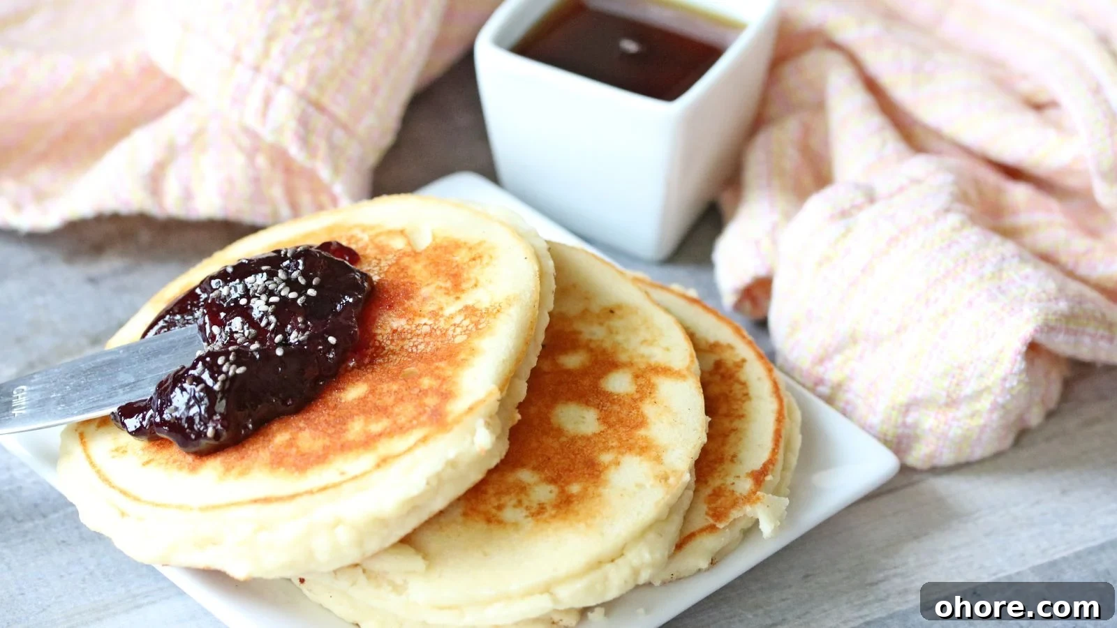 Three yogurt pancakes on a white plate topped with jam. Syrup in a white container behind it and a light orange napkin