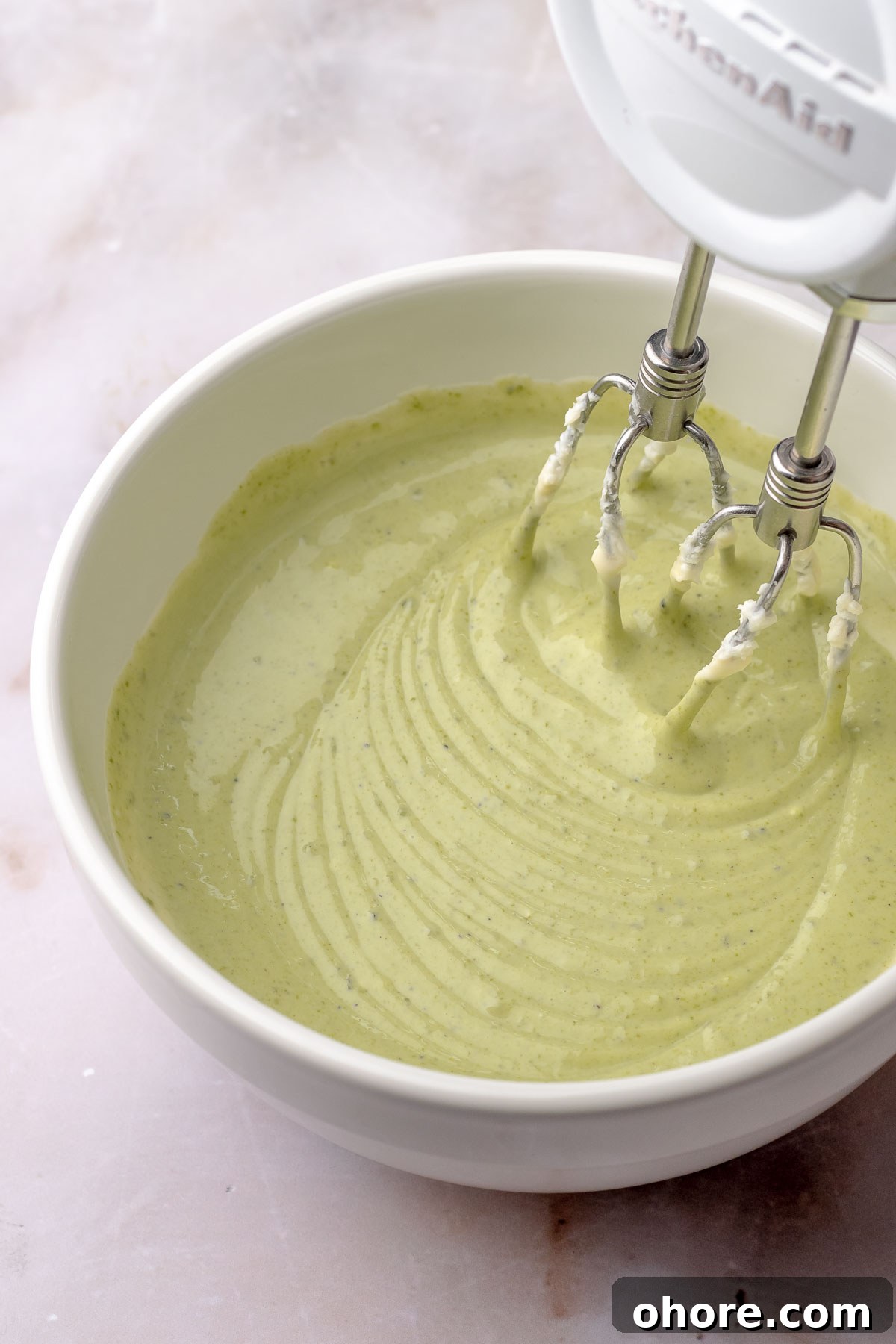 A hand mixes beats pistachio cheesecake batter in a bowl.