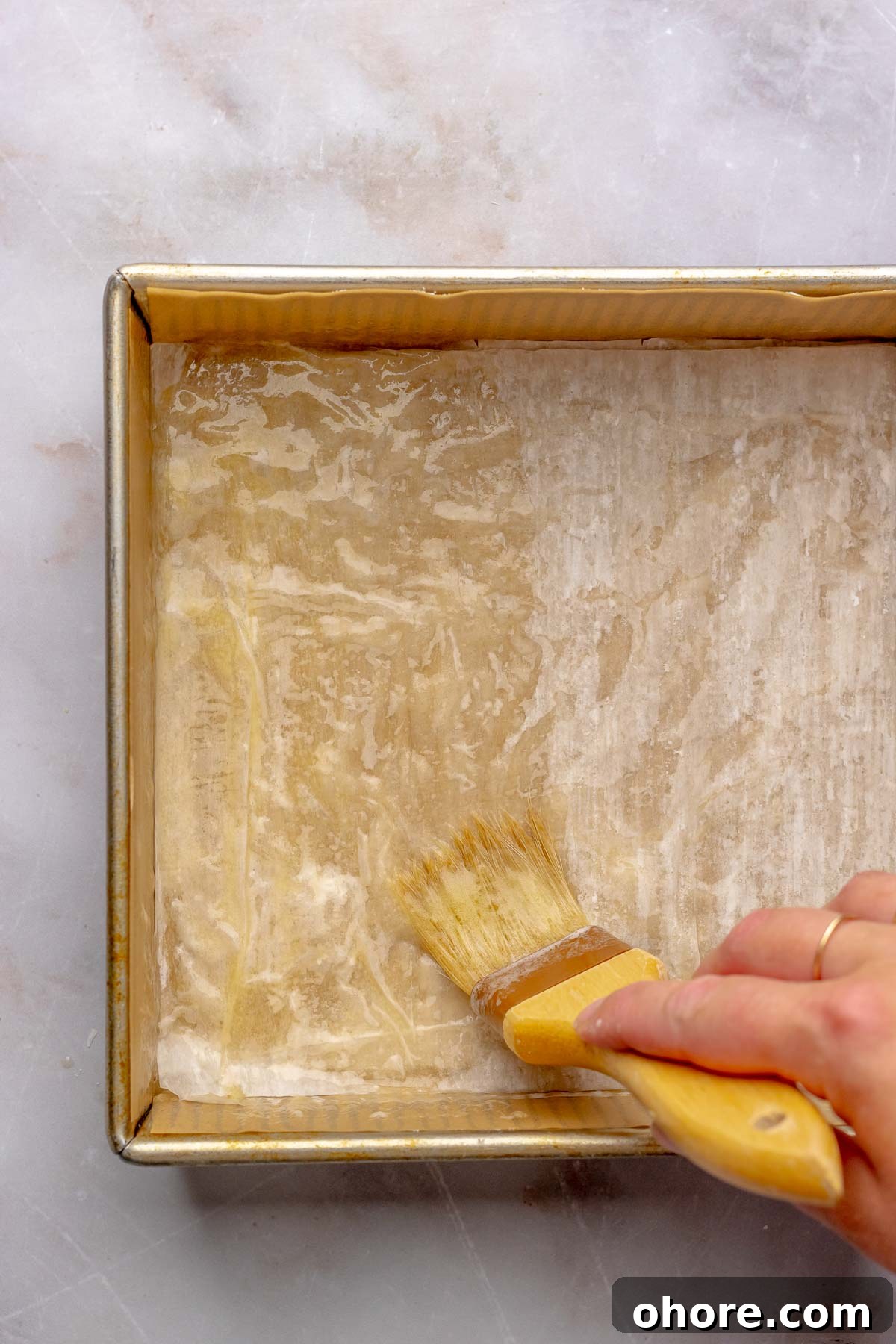 A brush swipes butter over phyllo pastry in a square pan.