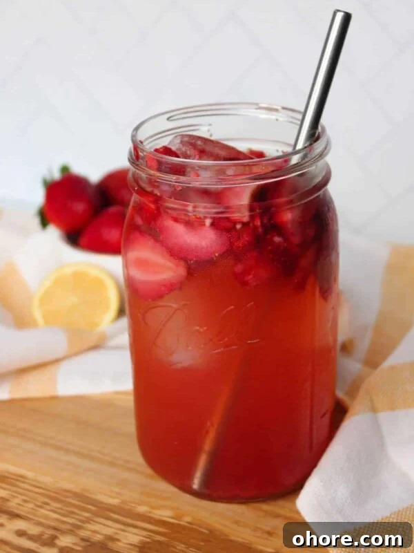 A mason jar full of iced strawberry green tea with a metal straw, strawberries and lemon visible in the background.