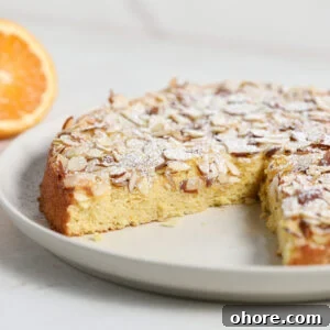 Moist Orange and Almond Cake with Zesty Citrus Glaze 2 A round Orange Almond Cake topped with sliced almonds and powdered sugar sits on a white plate. A slice has been removed, revealing its moist texture, and a halved orange is visible in the background.