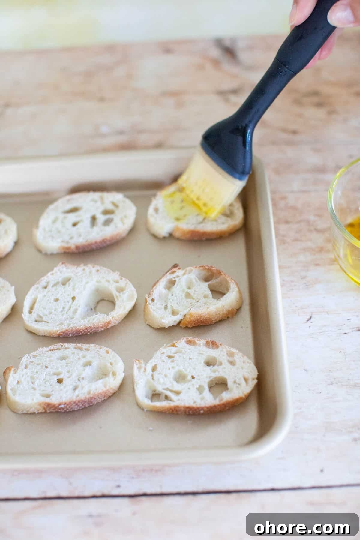 Slices of baguette on a baking sheet, being brushed in olive oil with a basting brush.
