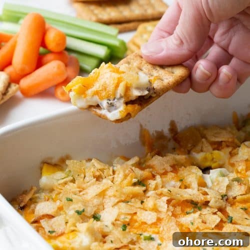 hand dipping a cracker into hot chicken salad topped with potato chips.