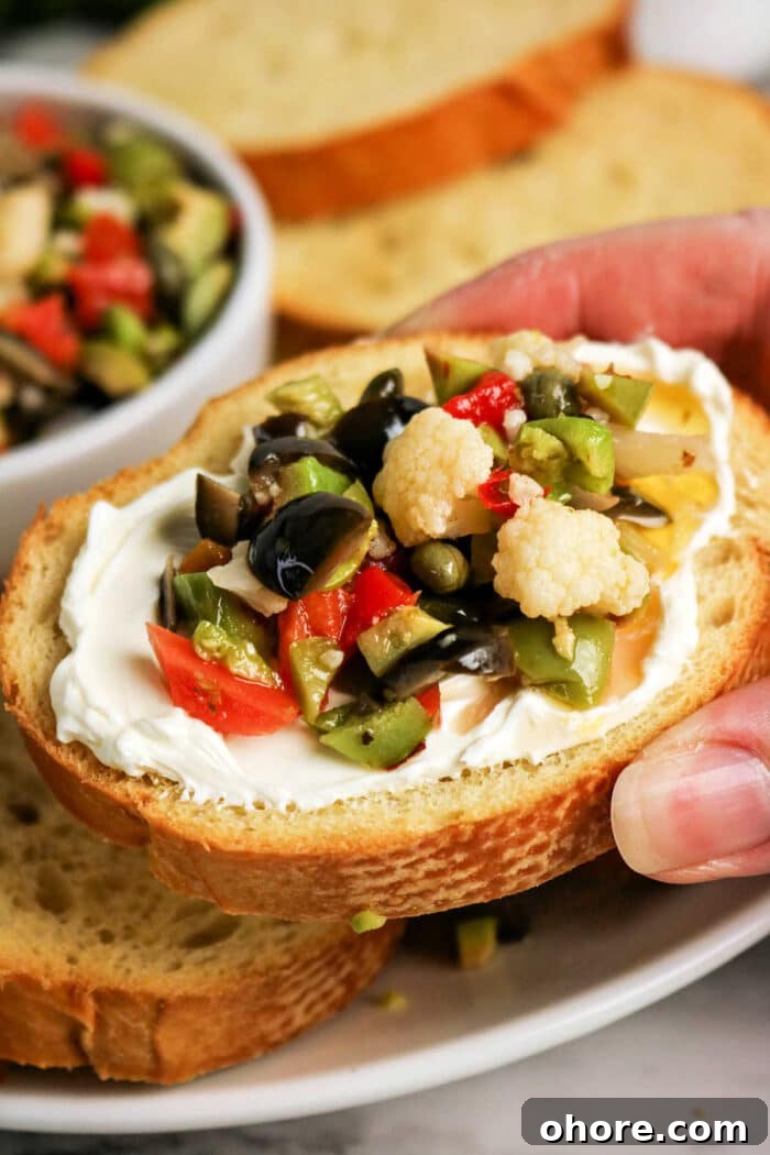 Authentic Muffuletta Olive Salad Recipe for Classic Sandwiches 2 A hand holds a slice of bread topped with cream cheese and a vibrant Olive Salad, featuring chopped olives, cauliflower, and peppers—a perfect bite inspired by the classic Muffuletta recipe.