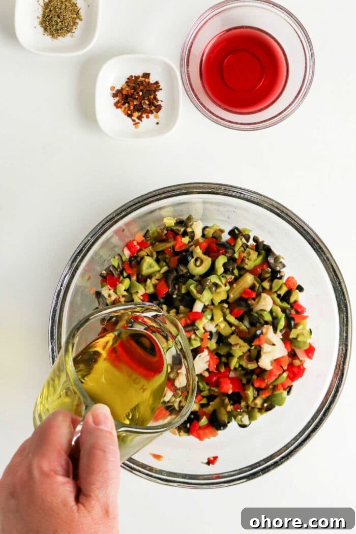 Authentic Muffuletta Olive Salad Recipe for Classic Sandwiches 4 A hand pours olive oil into a bowl of chopped vegetables for a classic Olive Salad, with spices and red vinegar nearby—perfect for an authentic Muffuletta recipe.