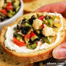 Authentic Muffuletta Olive Salad Recipe for Classic Sandwiches 6 A hand holds a slice of bread topped with cream cheese and a vibrant Olive Salad, featuring chopped olives, cauliflower, and peppers—a perfect bite inspired by the classic Muffuletta recipe.