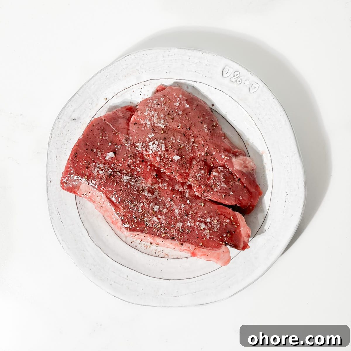 Raw steak on a plate that's been seasoned with salt and pepper.