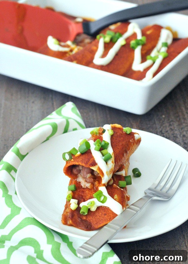 Bean and Rice Enchiladas one Bean and Cauliflower Rice Enchilada stacked on a second one, on a white plate next to a green and white cloth napkin, and a full pan of red sauce enchiladas in the background.