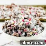 Valentine’s Puppy Chow: Festive Chocolate-Covered Snack Mix Recipe 15 Valentine's puppy chow made with pink m and m's and white chocolate chips in a white serving dish, with a tray of puppy chow in the background.