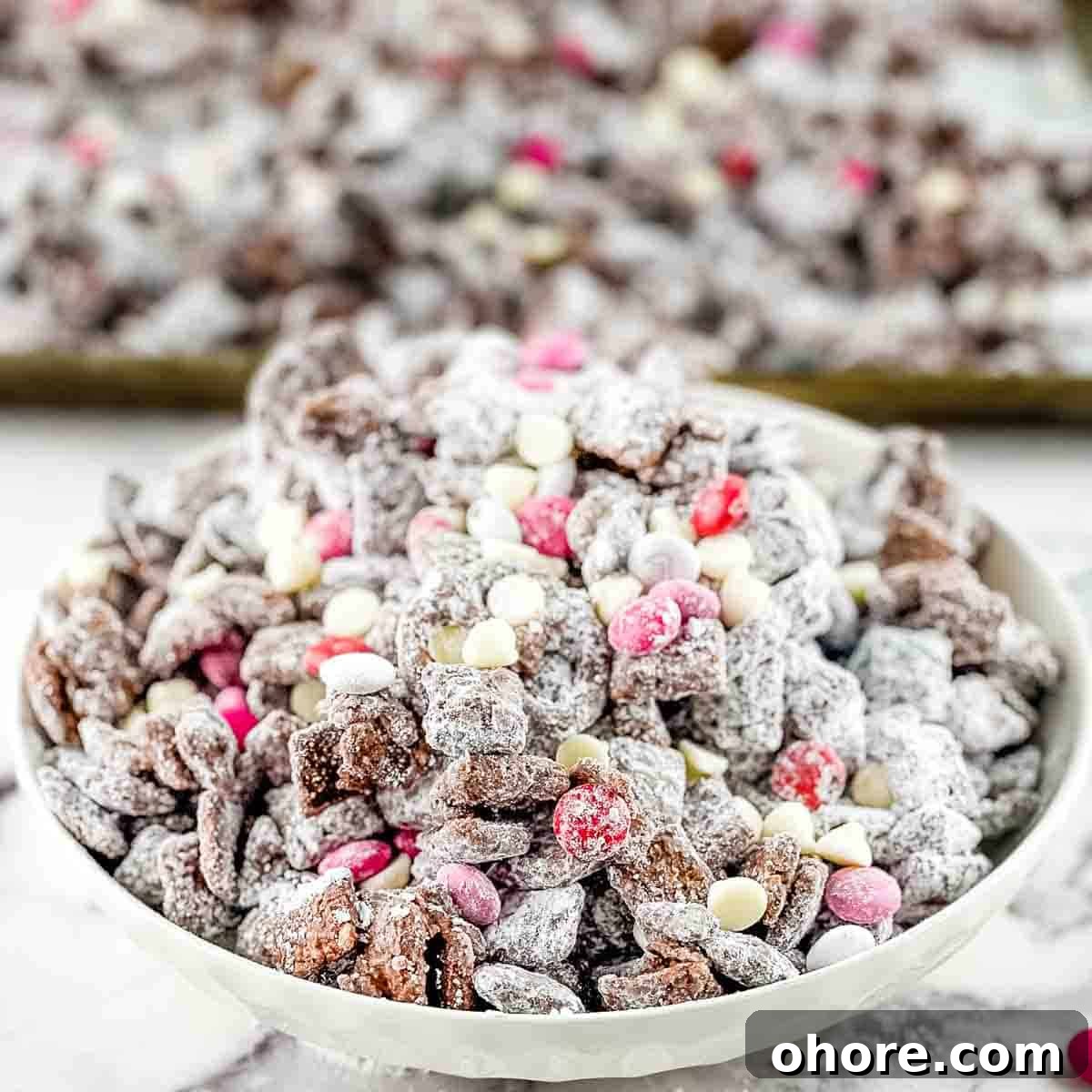 Valentine’s Puppy Chow: Festive Chocolate-Covered Snack Mix Recipe 9 The prepared puppy chow in a white serving bowl, with a baking sheet full of it in the background.