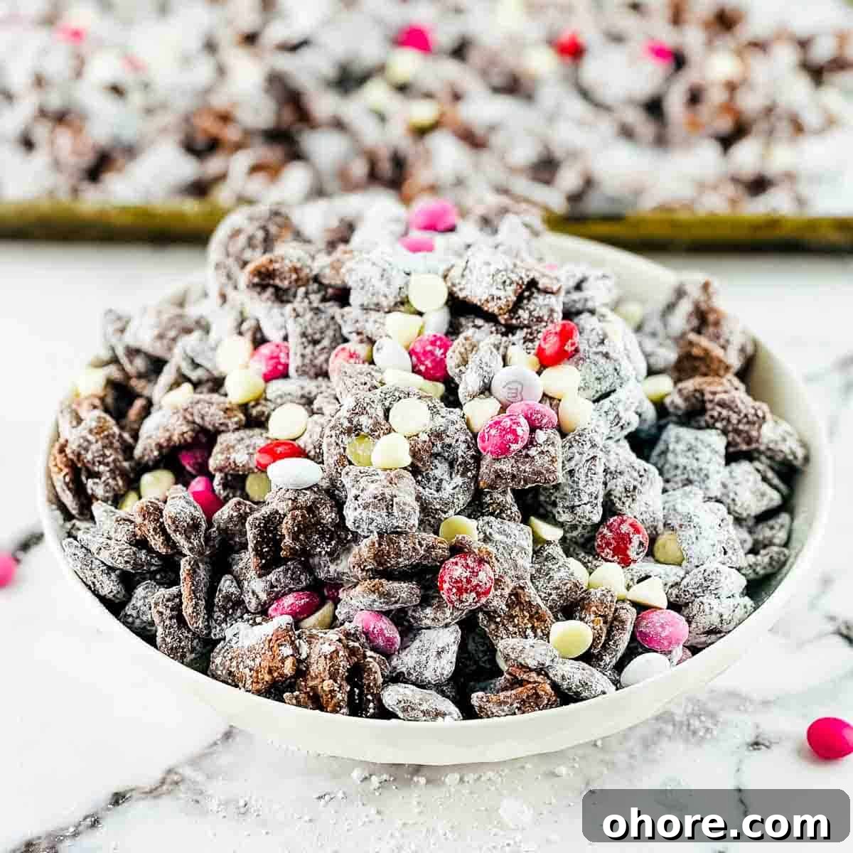 Valentine’s Puppy Chow: Festive Chocolate-Covered Snack Mix Recipe 10 Valentine's puppy chow made with pink m and m's and white chocolate chips in a white serving dish, with a tray of puppy chow in the background.