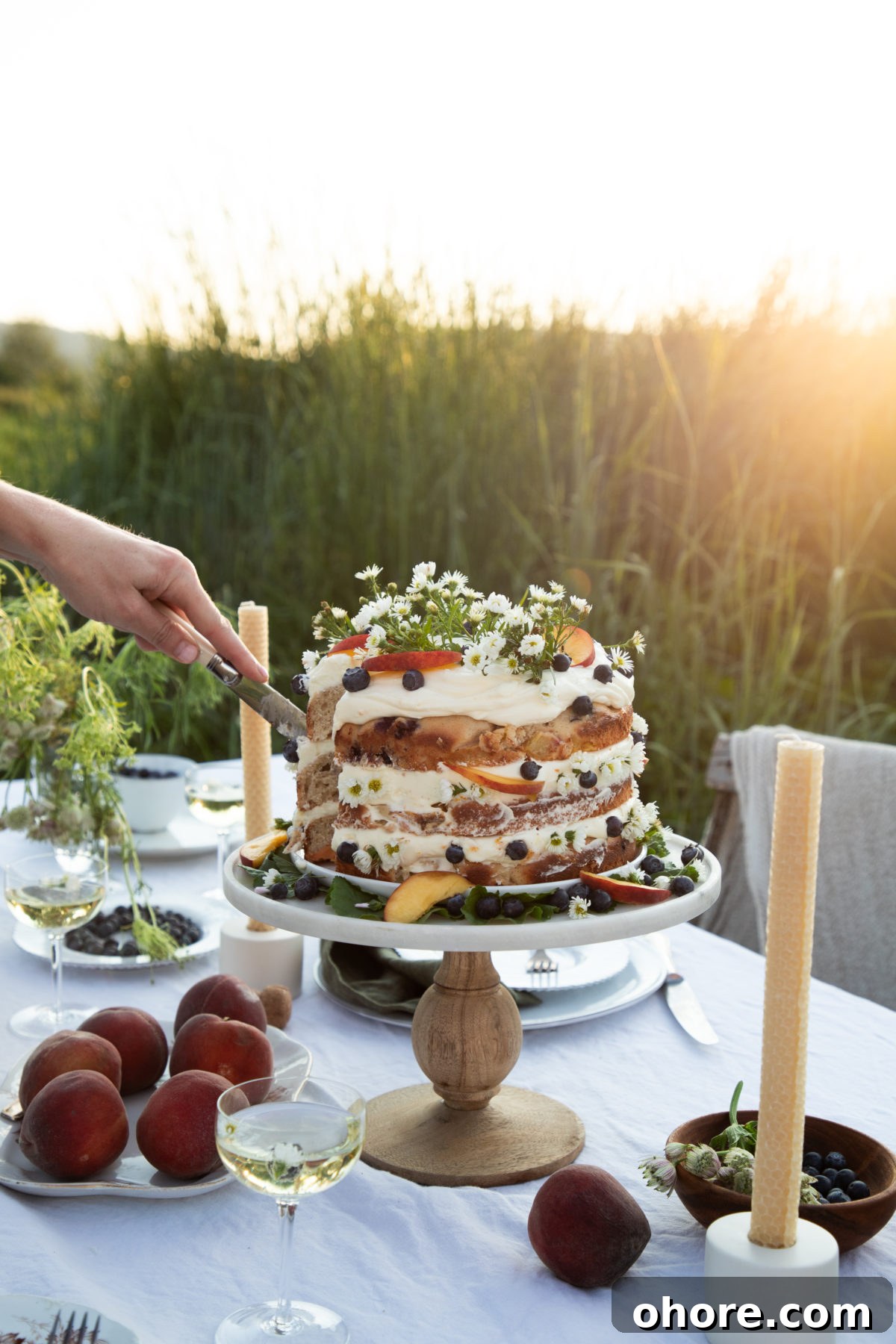 Ultimate Homemade Peach Cake Recipe for Moist, Fruity Perfection 3 a woman's hand cutting into a cake that's topped with peaches, blueberries, and camomile flowers on a white cloth covered table in a field with peaches and candlesticks on the table as well.