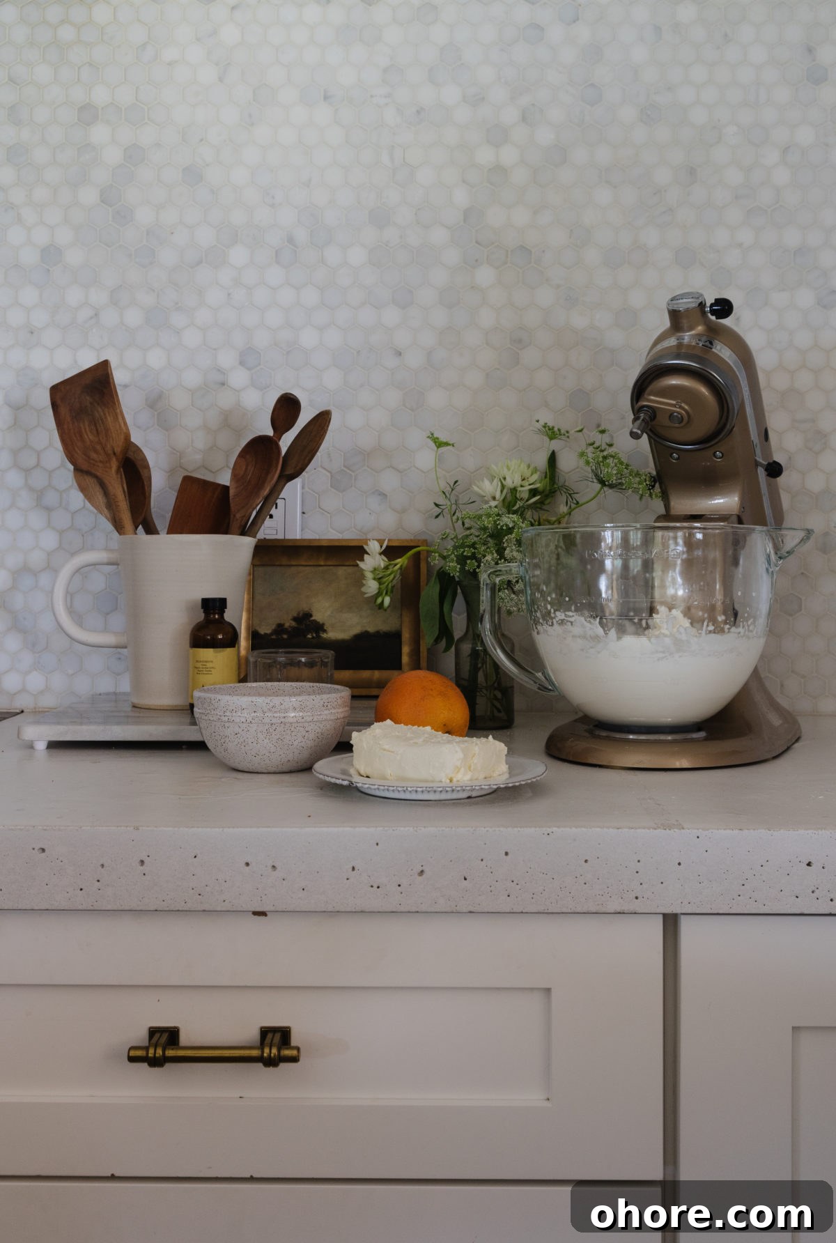Ultimate Homemade Peach Cake Recipe for Moist, Fruity Perfection 5 white kitchen with marble penny round backsplash and white concrete countertop with a white jar filled with wooden utensils, artwork, flowers, and the ingredients for cream cheese frosting next to a gold kitchen aid mixer.