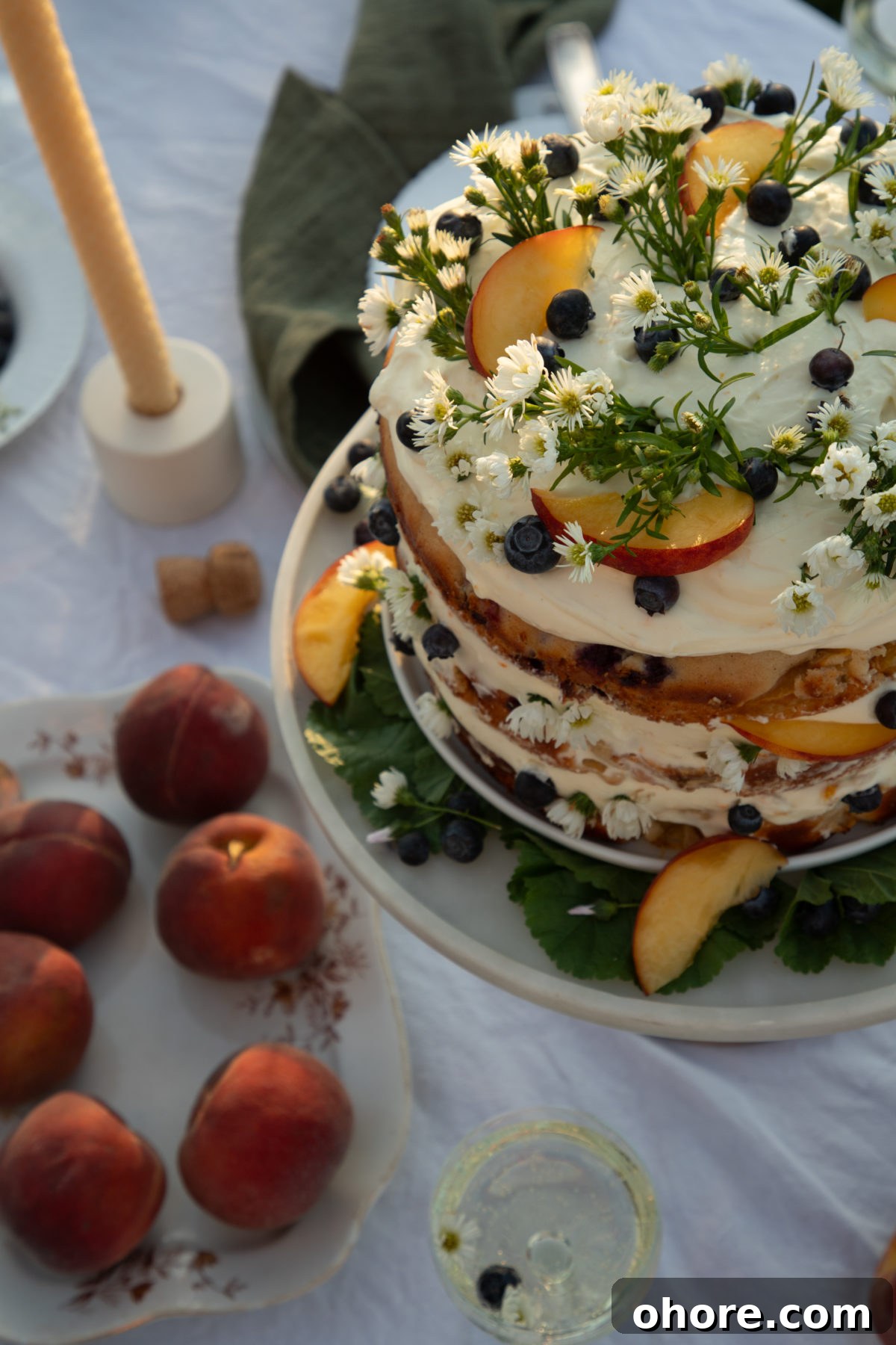 Ultimate Homemade Peach Cake Recipe for Moist, Fruity Perfection 7 a close up photo of a a cake with whipped cream cheese frosting topped with peach slices, blueberries, and camomile flowers with peaches on the table in front of the cake