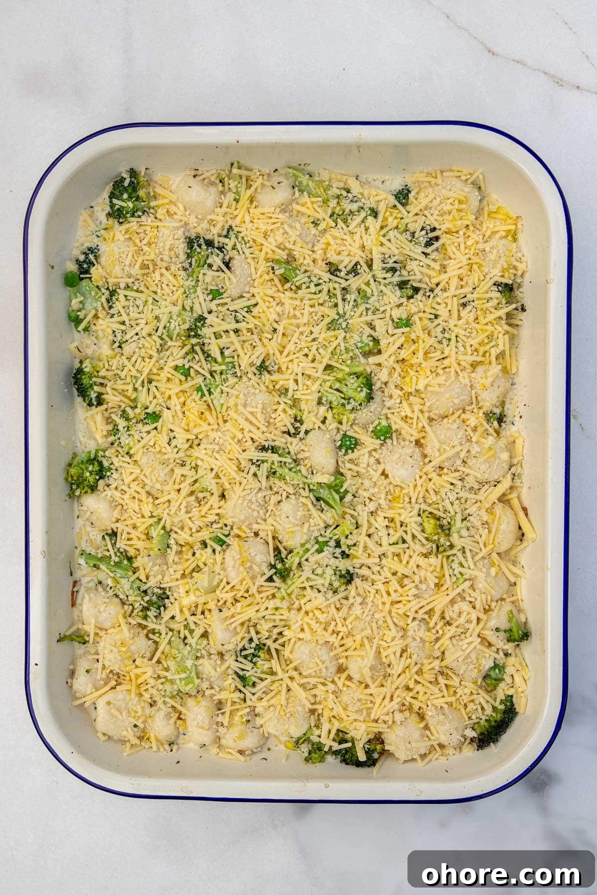 One-Pan Baked Creamy Broccoli Gnocchi Recipe 6 Overhead view of a white baking dish with a blue rim with broccoli gnocchi with cheese on top ready to be baked.