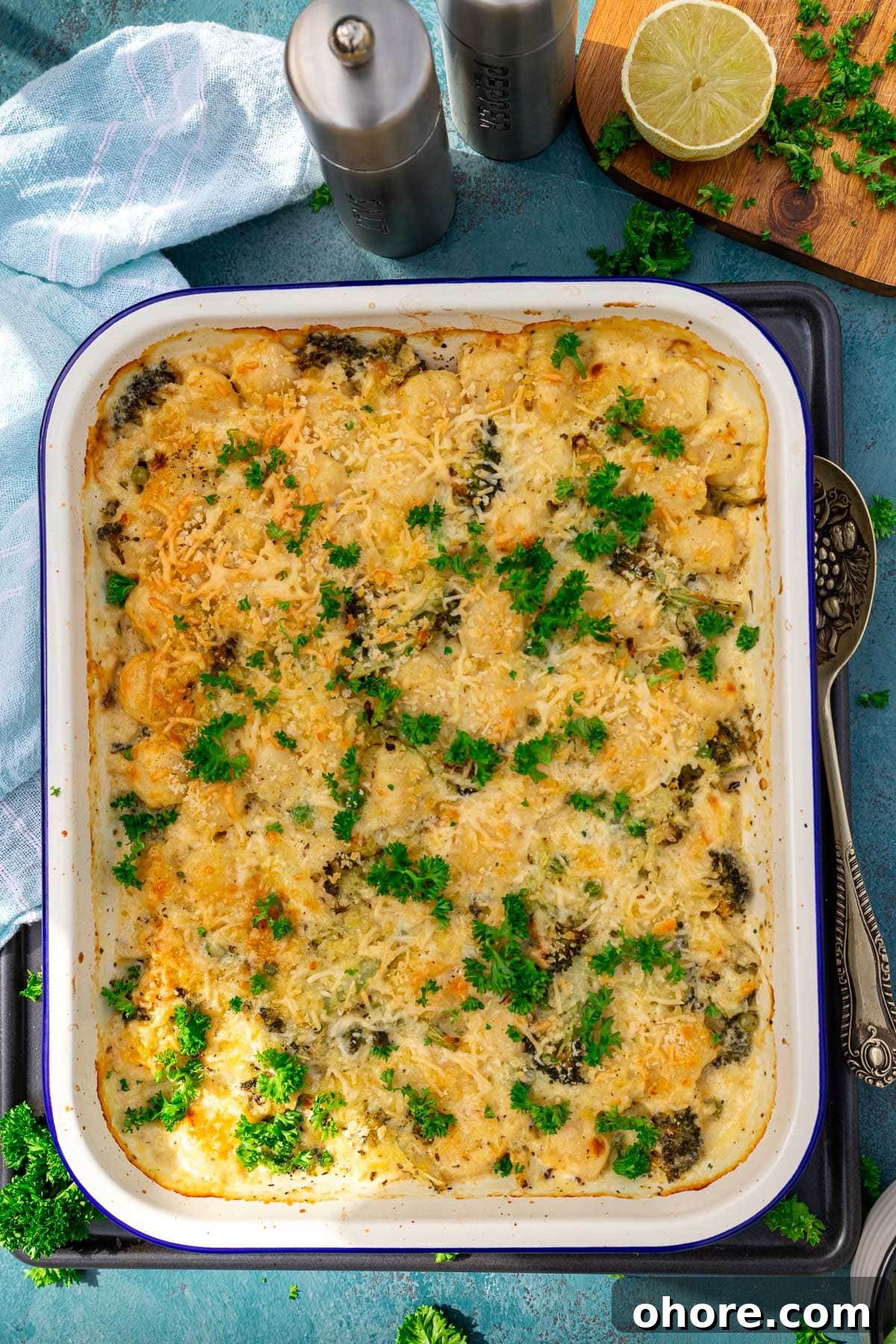 One-Pan Baked Creamy Broccoli Gnocchi Recipe 7 Overhead view of a white baking dish of baked broccoli gnocchi with a decorative spoon, pale blue tea towel, and ingredients at the side.