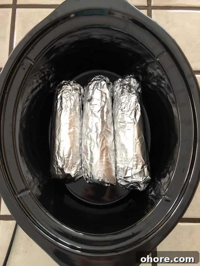 Three ears of corn on the cob wrapped in aluminum foil in black crock pot.