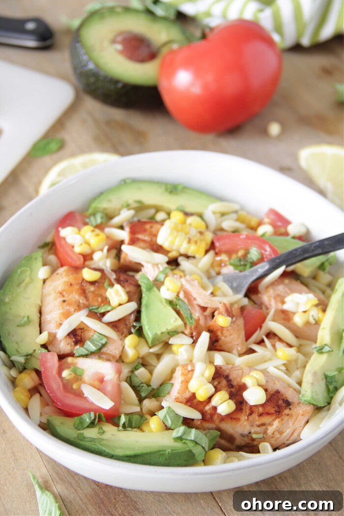 Grilled salmon in a white bowl over orzo pasta, sliced avocado and tomatoes, and grilled corn with a fork