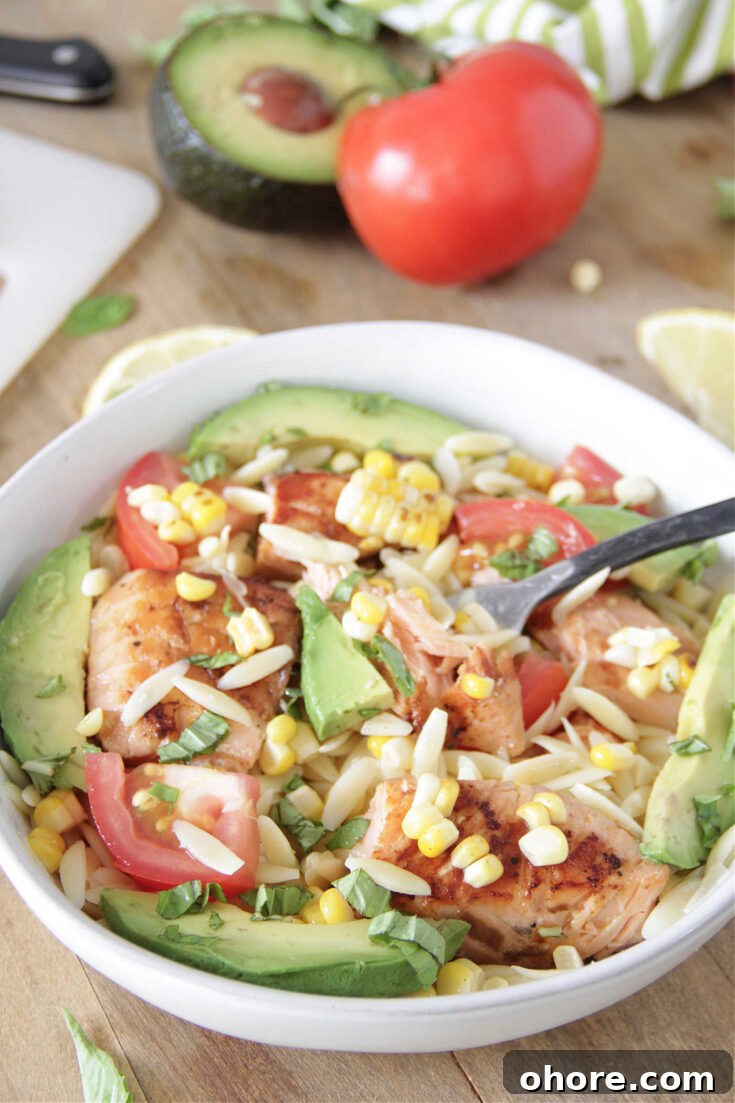 Grilled salmon in a white bowl over orzo pasta, sliced avocado and tomatoes, and grilled corn with a fork