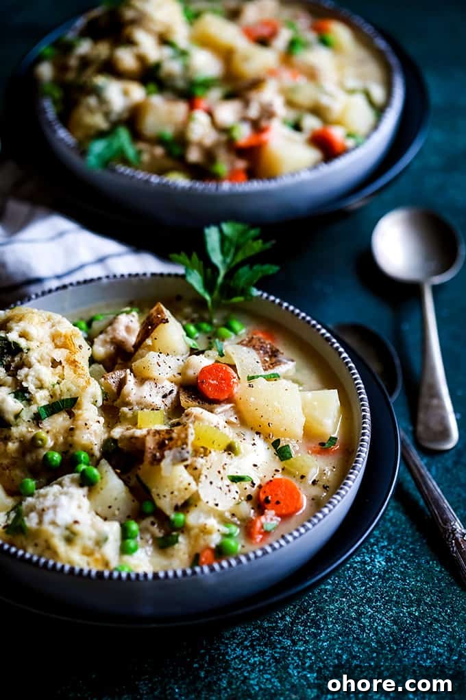 Easy Chicken and Dumpling Soup Two bowls of Easy Chicken and Dumpling Soup alongside two spoons and a striped napkin rest on a green speckled surface.