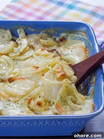 cooked scalloped potatoes without cheese in a blue casserole dish with a wooden spoon and a checkered tea towel in the background