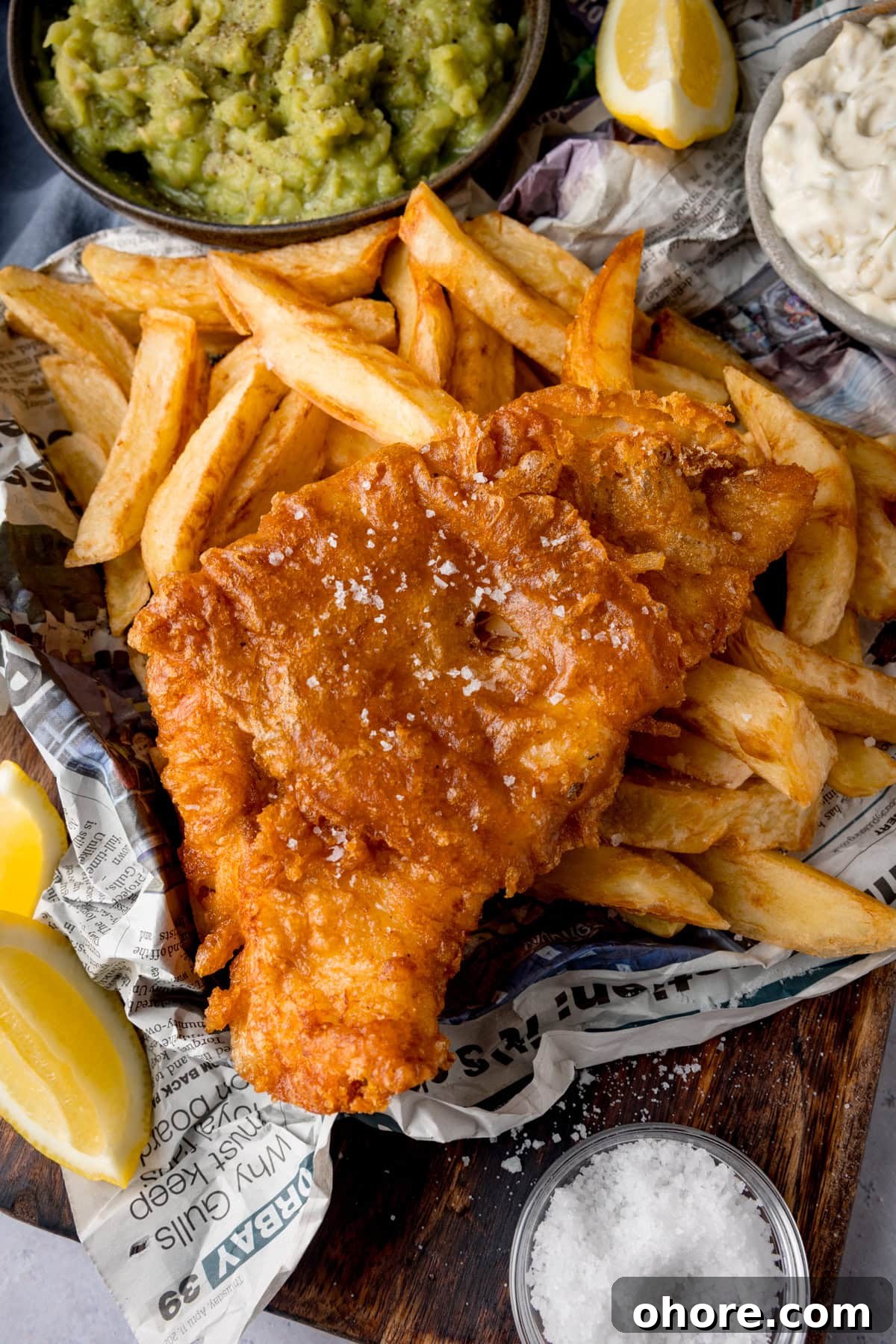 A tall, overhead shot of beer-battered fish. There is one beer-battered fish fillet, topped with flakes of salt, on a bed of chips. The food is on top of a newspaper. To the left of the fish and chips are two wedges of lemon. In the top left of the image, there is a black bowl of mushy peas, with another wedge of lemon to the right of it. In the top right of the image, there is a grey dish of tartare sauce. This is all on a wooden board. On the bottom right of the wooden board, there is a clear dish of sea salt. This is all placed on a white marble background.