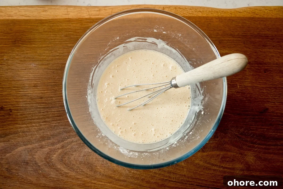 A wide, overhead shot of the finished beer batter. The batter is in a clear, glass bowl with a whisk wooden handle in the batter. All this is laid on a wooden board.