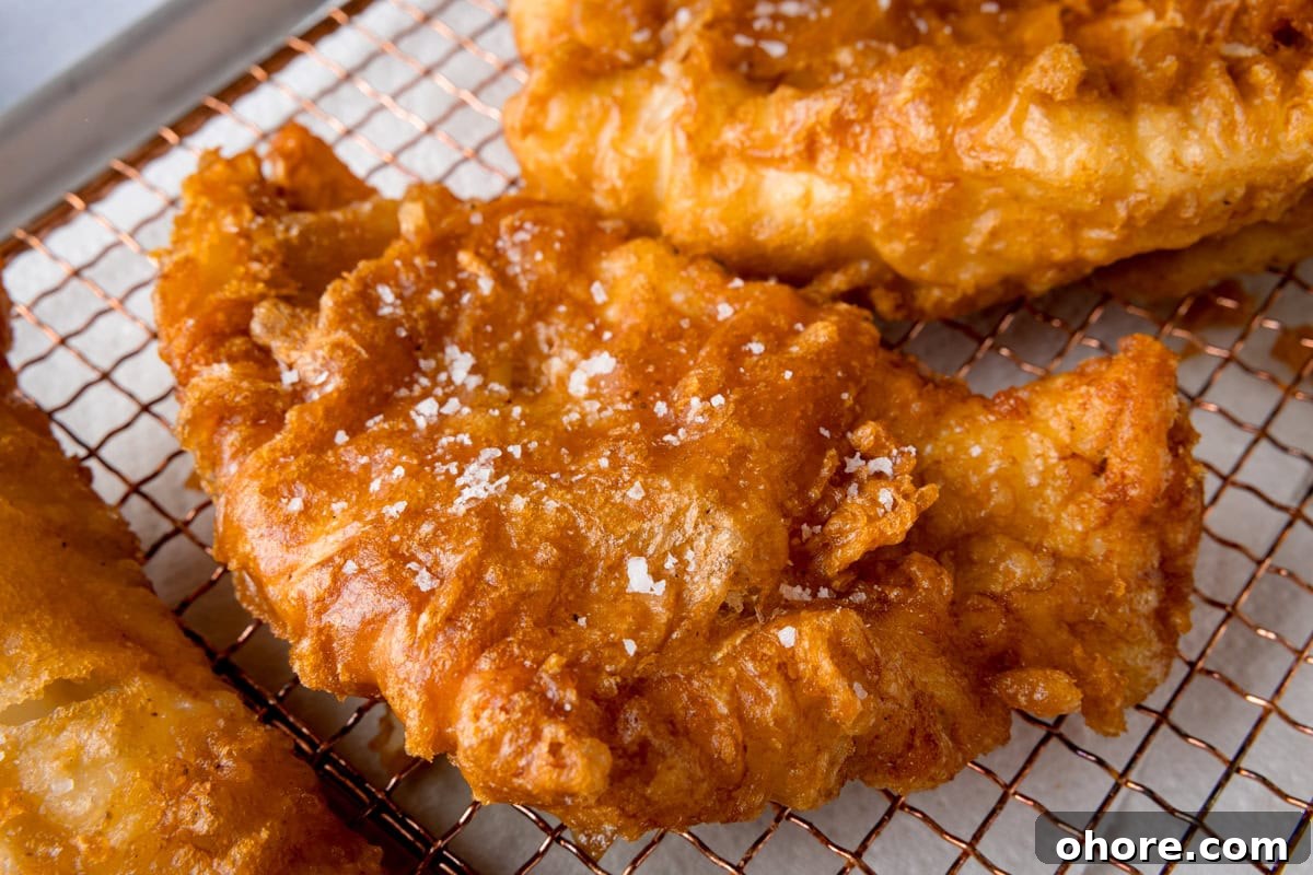A wide, close-up shot of three fish fillets, battered in the beer batter. There are three golden battered fish fillets laid side by side on a copper wire rack. They are topped with flakes of salt. The wire rack is on a white marble background.